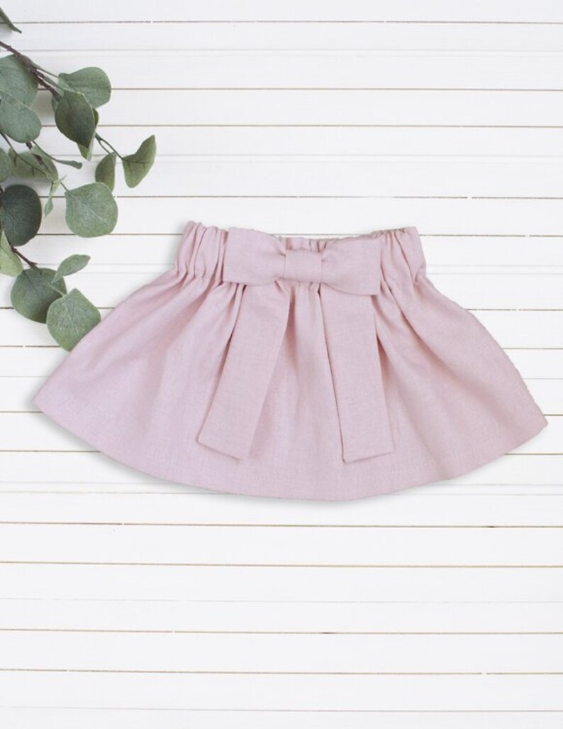 May include: A pale pink skirt with a large bow at the waist. The skirt is made of a soft, lightweight fabric.