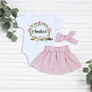 K&ouml;nnte beinhalten: Ein Baby-M&auml;dchen-Outfit-Set mit einem wei&szlig;en Strampler mit einem Blumenkranz mit dem Namen "Amber" in der Mitte, einem rosa Rock mit Schleife und einem rosa Stirnband mit Schleife.