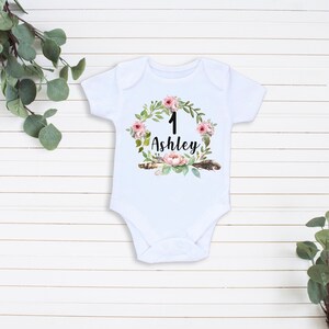 May include: White baby onesie with a floral wreath design. The wreath has pink flowers, green leaves, and feathers. The number "1" and the name "Ashley" are in the center of the wreath.