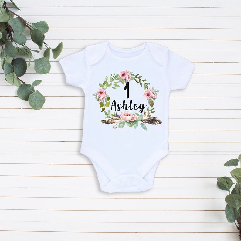 May include: White baby onesie with a floral wreath design. The wreath has pink flowers, green leaves, and feathers. The number "1" and the name "Ashley" are in the center of the wreath.