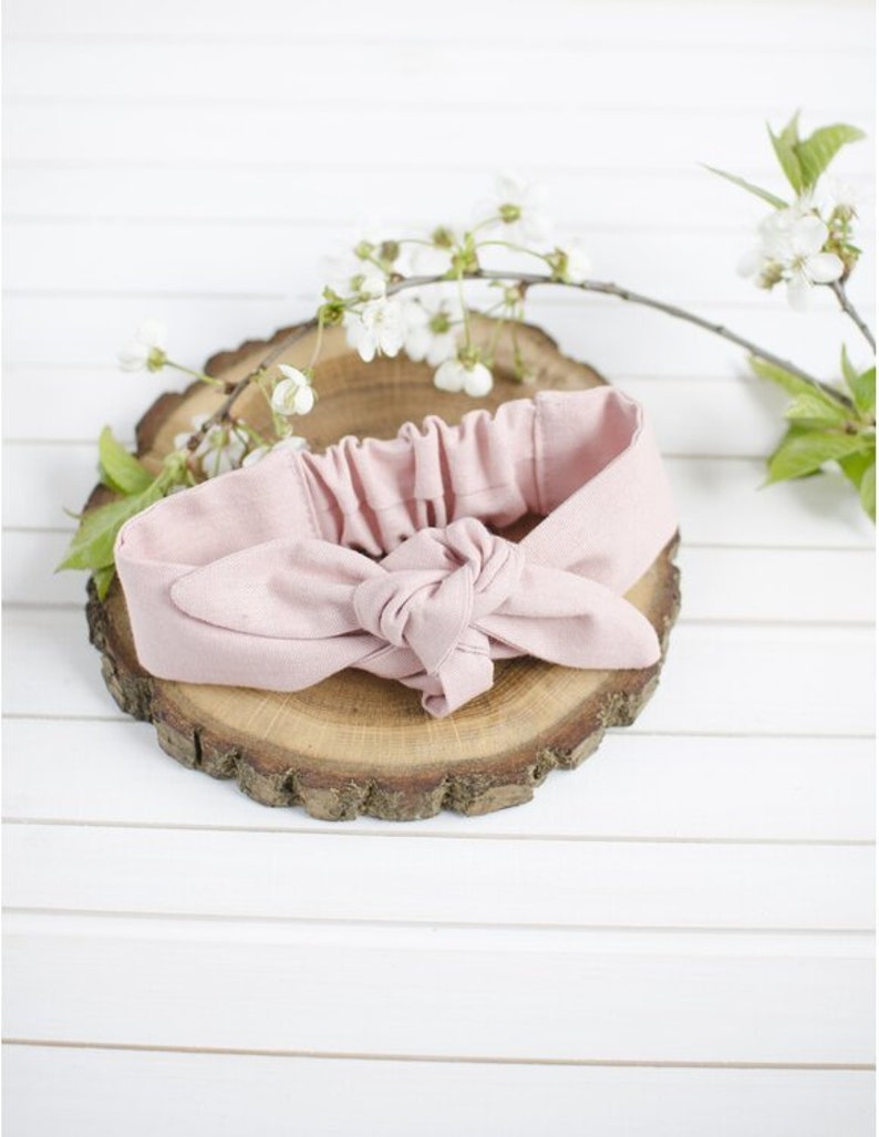 K&ouml;nnte beinhalten: Ein rosa Stoff-Haarband, das zu einem Knoten gebunden ist. Das Haarband liegt auf einer Holzscheibe.