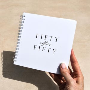 white spiral bound book with “Fifty after Fifty” on the cover behind held by a hand against a beige background