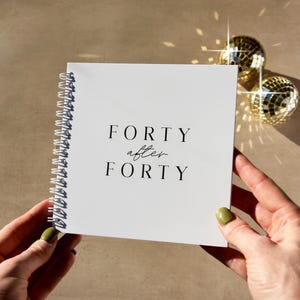 May include: A square white spiral-bound notebook with black text on the front cover that reads FORTY after FORTY. The notebook features a white metal coil binding along the left side and is held against a neutral background with two disco balls.