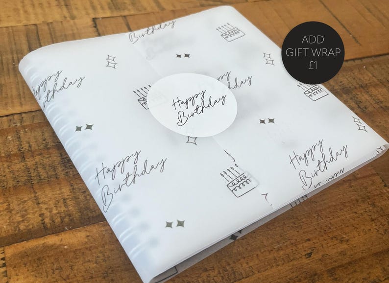 Op de afbeelding: Wit tissuepapier met een zwart-wit patroon van sterren, taarten en de woorden "Happy Birthday". Een witte ronde sticker met de woorden "Happy Birthday" zit op het tissuepapier. De tekst "ADD GIFT WRAP &pound;1" staat op een zwarte ronde sticker.