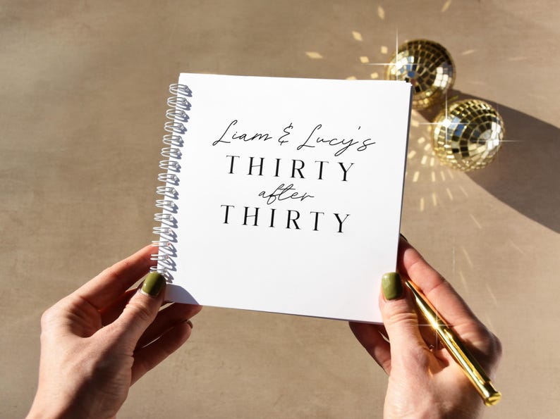 May include: A white spiral-bound notebook with the text "Liam & Lucy's THIRTY after THIRTY" in black script. The notebook is held by hands with green nail polish. Two disco ball ornaments and a gold pen are also visible.