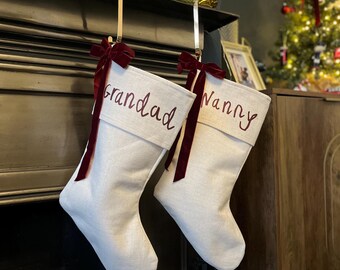 Op de afbeelding: Twee gebroken witte kerstsokken van stof die aan metalen haken hangen. Elke sok is aan de bovenkant voorzien van een donkerrode fluwelen strik en geborduurd met donkerrode tekst. Op de ene sok staat Grandad en op de andere staat Nanny.