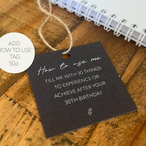 May include: A dark gray tag with the text "How to use me" and instructions to fill it with 30 things to experience or achieve after your 30th birthday. The tag is attached to a natural twine string.