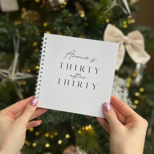 May include: A white spiral-bound notebook with the text "Annie's Thirty After Thirty" in black script. The notebook is held in front of a blurred background of a Christmas tree with a bow.