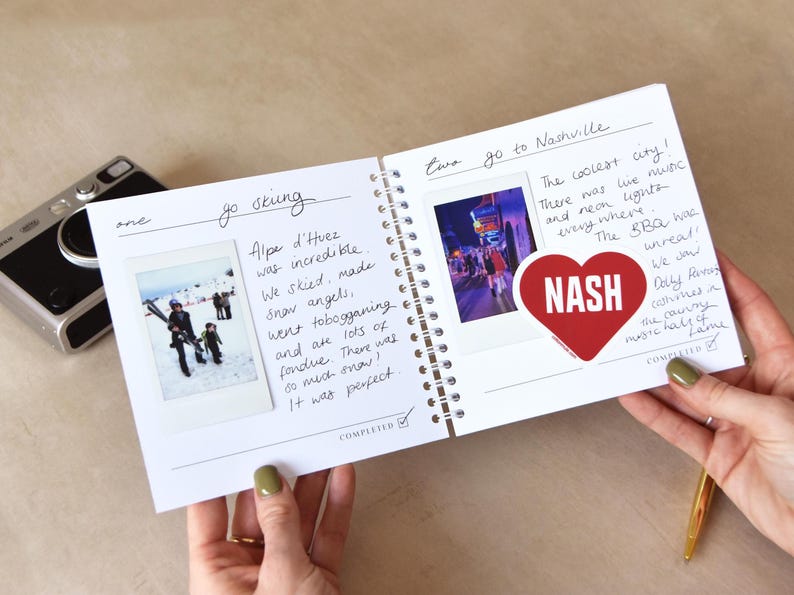 May include: A spiral-bound travel journal with handwritten notes and photos. One page features a photo of people skiing with the text "go skiing" and "Alpe d'Huez". The other page has a photo with the text "go to Nashville" and a heart-shaped sticker that says "NASH".
