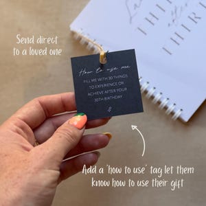 May include: A square black paper tag with white text that reads How to use me, FILL ME WITH 30 THINGS TO EXPERIENCE OR ACHIEVE AFTER YOUR 30TH BIRTHDAY, and a small sparkle icon. The tag is attached by a thin string to a white spiral-bound notebook.