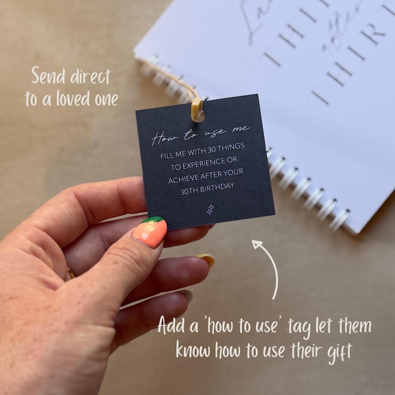 May include: A square black paper tag with white text that reads How to use me, FILL ME WITH 30 THINGS TO EXPERIENCE OR ACHIEVE AFTER YOUR 30TH BIRTHDAY, and a small sparkle icon. The tag is attached by a thin string to a white spiral-bound notebook.