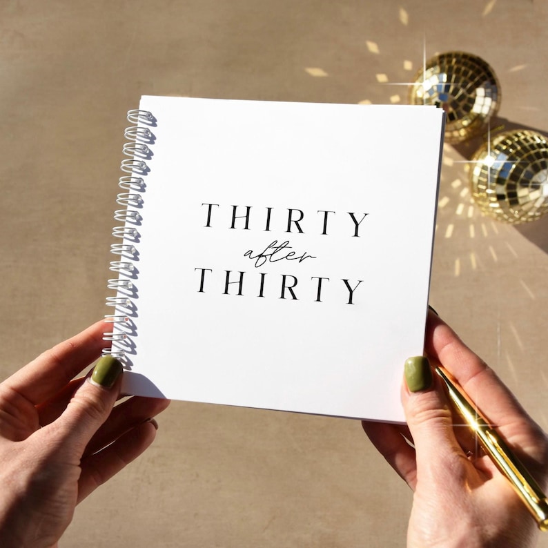 May include: A white spiral-bound notebook with the words "THIRTY after THIRTY" printed in black. The notebook is held by hands with green nail polish, and a gold pen rests on the right side. Two gold disco balls are in the background.
