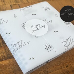 May include: White gift wrap with a repeating pattern of birthday cakes, stars, and the words "Happy Birthday" in a handwritten font. A round sticker with "Happy Birthday" is on top.