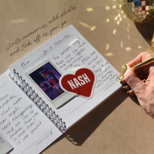 May include: A spiral-bound journal with handwritten notes and a photo. A red heart-shaped sticker with "NASH" is on the page. The text "Write memories, add photos and tick off as you go..." is written at the top.