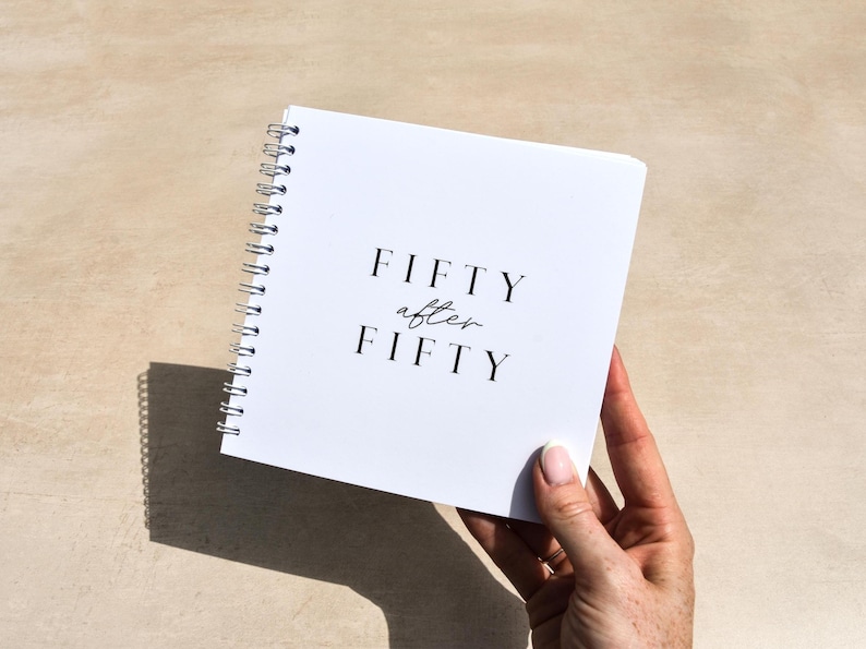 white spiral bound book with “Fifty after Fifty” on the cover behind held by a hand against a beige background