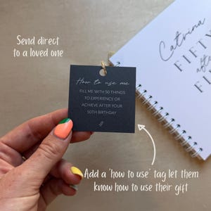 Może przedstawiać: Kwadratowa czarna zawieszka z białym napisem How to use me, FILL ME WITH 50 THINGS TO EXPERIENCE OR ACHIEVE AFTER YOUR 50TH BIRTHDAY, umieszczona obok białego notatnika spiralnego z napisem Catrina FIFTY after FIFTY.