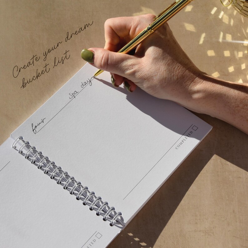 May include: A gold pen writing on a white spiral-bound notebook. The notebook is open to a blank page with the words "Spa day" and "four" written on it. The text "Create your dream bucket list" is written in cursive.