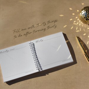 May include: A spiral-bound notebook with the text "Fill me with thirty things to do after turning thirty." The open pages display the words "twenty-nine" and "thirty." A gold pen and disco ball are also visible.