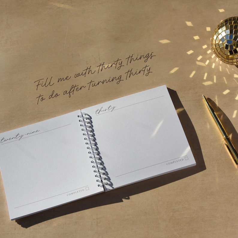 May include: A spiral-bound notebook with the text "Fill me with thirty things to do after turning thirty." The open pages display the words "twenty-nine" and "thirty." A gold pen and disco ball are also visible.