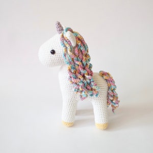 White crocheted amigurumi unicorn with a pastel rainbow mane, lilac horn and yellow hooves
