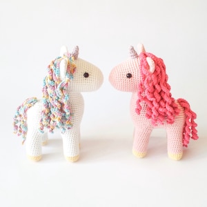 On the left, white crocheted amigurumi unicorn with a pastel rainbow mane, lilac horn and yellow hooves. On the right, pink unicorn with a bright pink mane, lilac horn and yellow hooves.