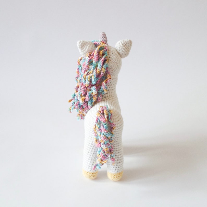 Back view of a white crocheted amigurumi unicorn with a pastel rainbow mane, lilac horn and yellow hooves