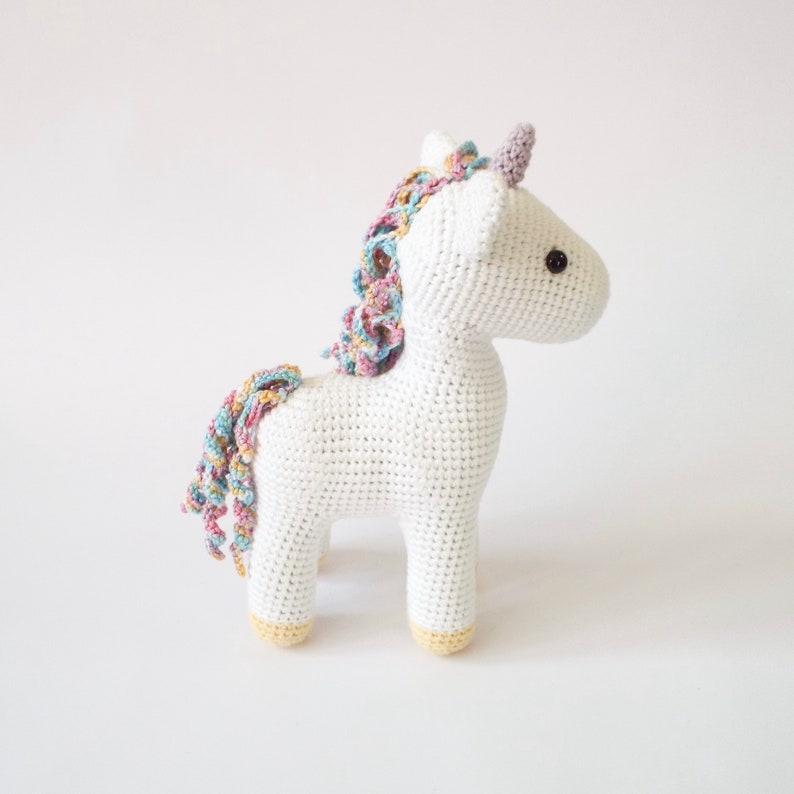 White crocheted amigurumi unicorn with a pastel rainbow mane, lilac horn and yellow hooves