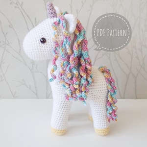 White crocheted unicorn with a pastel rainbow mane, lilac horn and yellow hooves in front of a background of silver trees