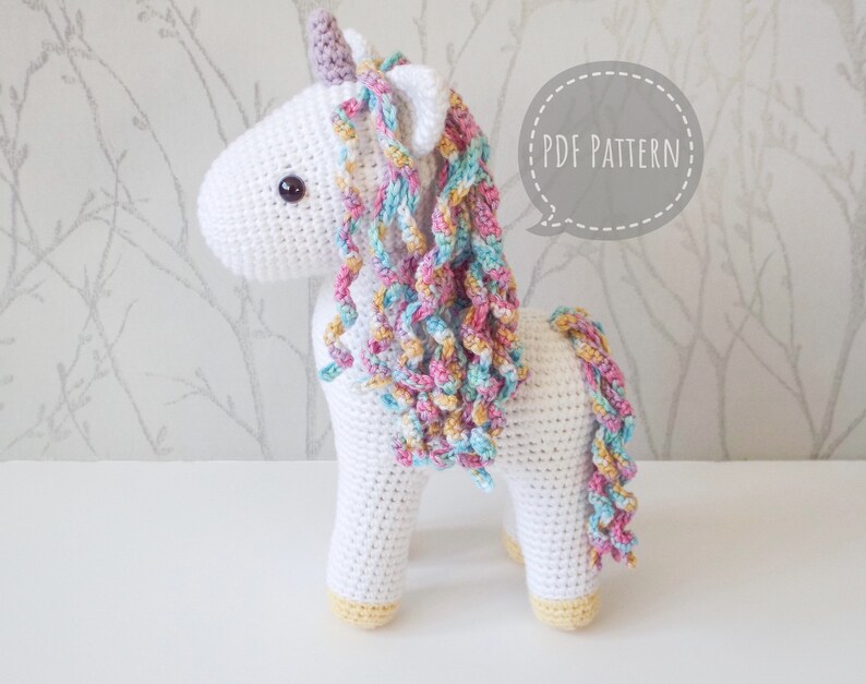 White crocheted unicorn with a pastel rainbow mane, lilac horn and yellow hooves in front of a background of silver trees