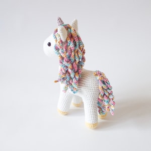 Back view of a white crocheted amigurumi unicorn with a pastel rainbow mane, lilac horn and yellow hooves