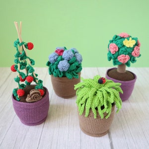 Puede incluir: Cuatro plantas en maceta de ganchillo, incluyendo una planta de tomate con bayas rojas, una planta de flores azules, una planta de flores rosas y una planta verde con una mariquita. Las plantas están en macetas marrones y moradas.