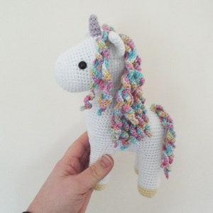 Hand holding a white crocheted amigurumi unicorn with a pastel rainbow mane, lilac horn and yellow hooves