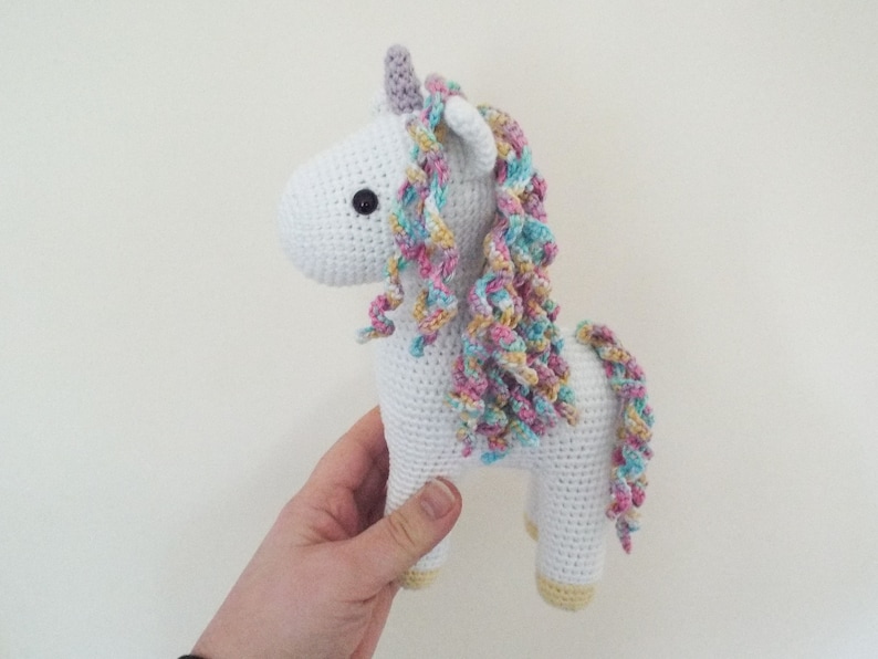 Hand holding a white crocheted amigurumi unicorn with a pastel rainbow mane, lilac horn and yellow hooves