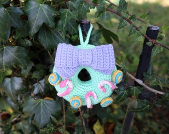 May include: A handmade, crocheted birdhouse in a whimsical design. The birdhouse is primarily mint green, adorned with a lavender bow and colorful candy-like embellishments. It hangs from a green loop against a backdrop of green foliage.