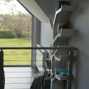 May include: White bookshelf shaped like a tree branch with books on the shelves. The bookshelf is mounted on a wall in a room with a large window and a radiator.