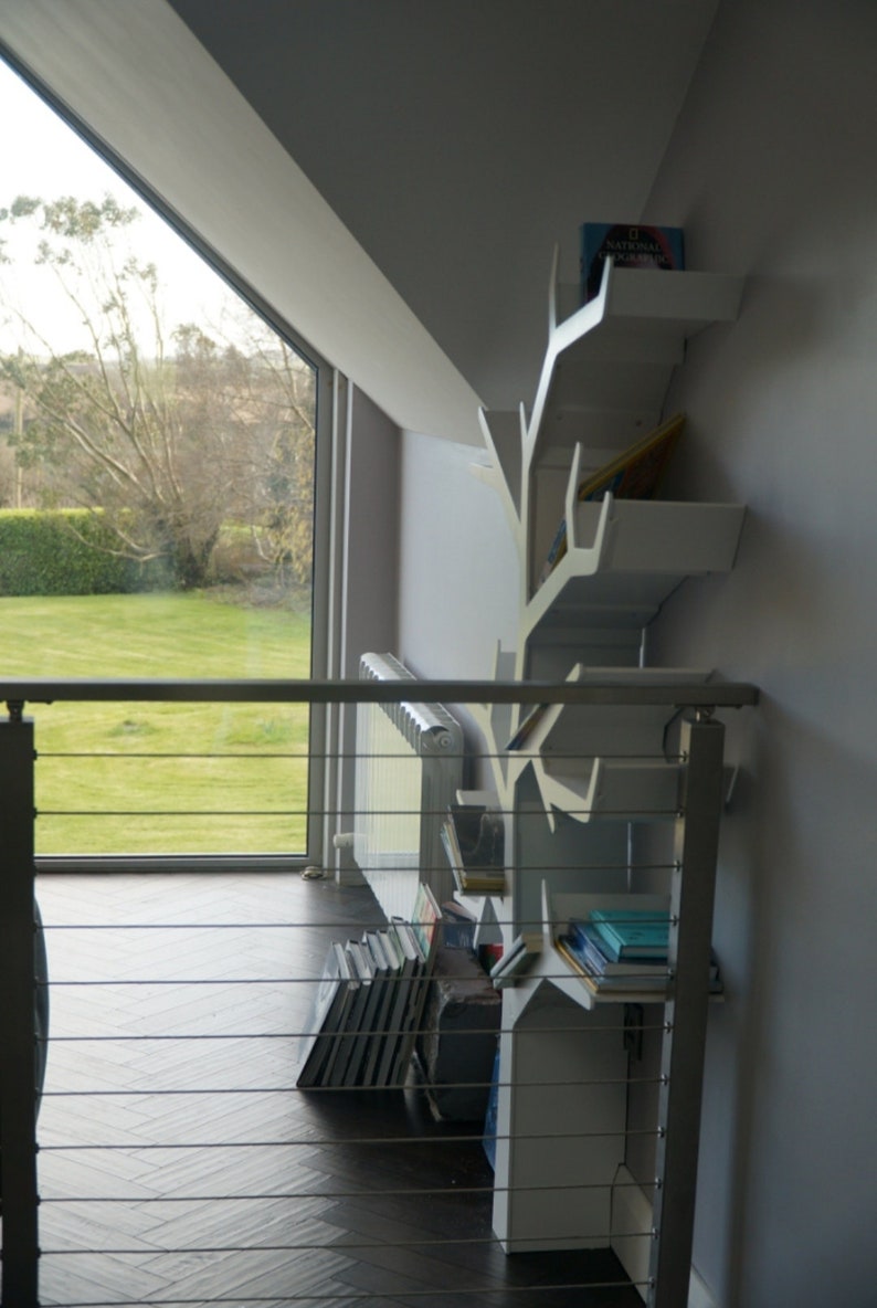 May include: White bookshelf shaped like a tree branch with books on the shelves. The bookshelf is mounted on a wall in a room with a large window and a radiator.
