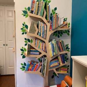 May include: A tree-shaped bookshelf made of light brown wood, mounted on a white wall. The bookshelf is filled with colorful children's books. Green leaf decals decorate the wall around the bookshelf, creating a whimsical reading nook.