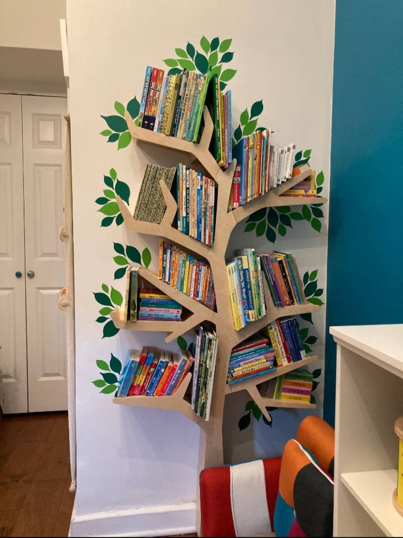 May include: A tree-shaped bookshelf made of light brown wood, mounted on a white wall. The bookshelf is filled with colorful children's books. Green leaf decals decorate the wall around the bookshelf, creating a whimsical reading nook.