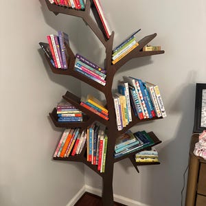 May include: A dark brown, tree-shaped bookshelf filled with colorful books. The bookshelf is made of wood and has multiple branches that serve as shelves. The books are of various sizes and colors, creating a visually appealing display. The bookshelf is placed in a corner.