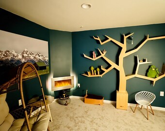 Puede incluir: Una habitación con paredes verde azulado presenta una gran estantería en forma de árbol, una chimenea moderna y una pintura de paisaje de montaña. También hay una silla de mimbre y una silla blanca con patas de madera. La habitación tiene un ambiente acogedor y acogedor.