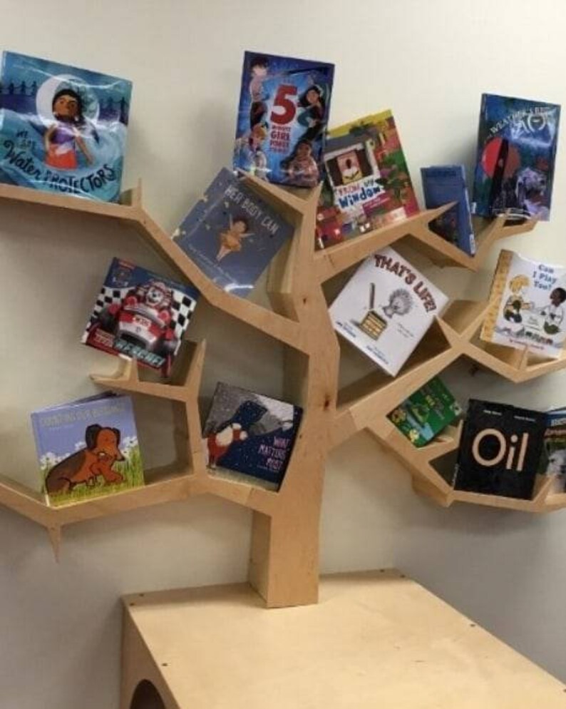 K&ouml;nnte beinhalten: Ein baumf&ouml;rmiges B&uuml;cherregal aus Holz mit verschiedenen Kinderb&uuml;chern. Die B&uuml;cher haben farbenfrohe Einb&auml;nde mit Titeln wie "Water Protectors" und "That's Life!". Das B&uuml;cherregal ist aus hellem Holz gefertigt und an einer Wand befestigt.