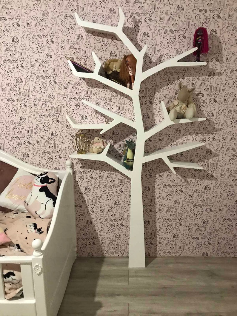 May include: White tree-shaped bookshelf with multiple shelves, decorated with stuffed animals and dolls. The bookshelf is against a pink wall with a black and white dog pattern.