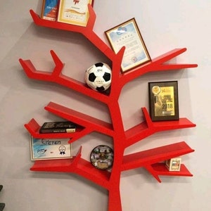 May include: Red tree-shaped bookshelf with multiple shelves and branches. The shelves are decorated with books, diplomas, a football, and other items.