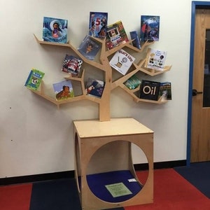 K&ouml;nnte beinhalten: Ein baumf&ouml;rmiges B&uuml;cherregal aus Holz mit mehreren Regalen, die verschiedene Kinderb&uuml;cher halten. Das B&uuml;cherregal hat eine quadratische Basis mit einer kreisf&ouml;rmigen &Ouml;ffnung. Die B&uuml;cher haben farbenfrohe Einb&auml;nde und Titel. Das B&uuml;cherregal steht an einer wei&szlig;en Wand.