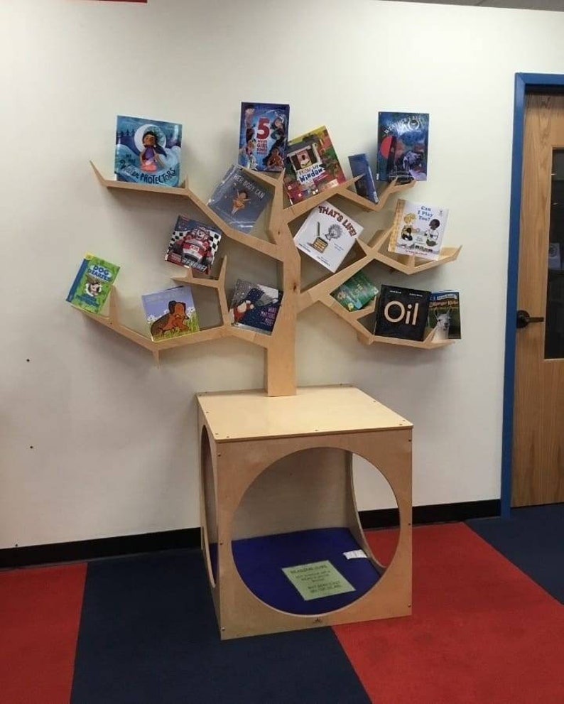K&ouml;nnte beinhalten: Ein baumf&ouml;rmiges B&uuml;cherregal aus Holz mit mehreren Regalen, die verschiedene Kinderb&uuml;cher halten. Das B&uuml;cherregal hat eine quadratische Basis mit einer kreisf&ouml;rmigen &Ouml;ffnung. Die B&uuml;cher haben farbenfrohe Einb&auml;nde und Titel. Das B&uuml;cherregal steht an einer wei&szlig;en Wand.