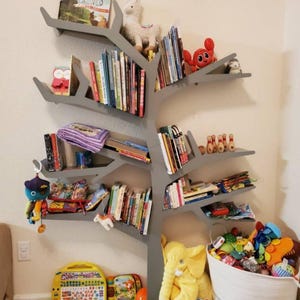 May include: A grey tree-shaped bookshelf with multiple branches and shelves filled with books and toys. A stuffed llama, elephant, and crab are on the shelves. A basket filled with toys is on the floor below the bookshelf.