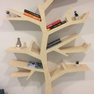 May include: A light wood bookshelf shaped like a tree with multiple branches and shelves. The shelves are filled with books and small potted plants.