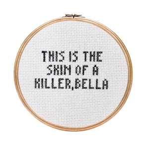 May include: A white embroidery hoop with the text "This is the skin of a killer, Bella" stitched in black thread.