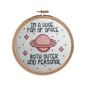 以下が含まれることがあります： ピンクの惑星と星が描かれた白い刺繍枠で、"私は宇宙の大ファンです。外側も内側も"というテキストが書かれています。