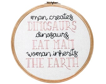 May include: A round wooden embroidery hoop with white fabric stretched across it. The fabric has a black and pink embroidered saying: "man creates DINOSAURS dinosaurs EAT MAN woman inherits THE EARTH"
